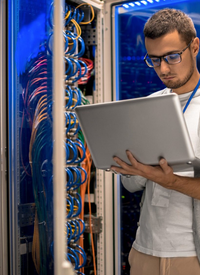 Portrait of modern young man holding laptop standing in server room working with supercomputer in blue light